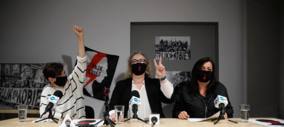 Trzy liderki Strajku Kobiet w czarnych maeczkach z błyskawicami pokazują znak V, siedząv przy stole z plakatami OSK