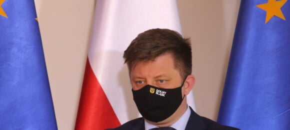 Michał Dworczyk w maseczce na twarzy na tle flag polskiej i unijnej