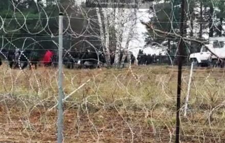 Migranci z plecakami są wyprowadzani znad granicy, widok przez zasieki z drutu żyletkowego