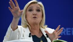 Kobieta z blond włosami macha rękami na trybunie, Marine Le Pen