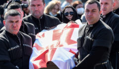 Mężczyźni w czarnych uroczystych strojach niosą trumnę na której jest gruzińska flaga