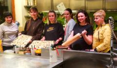 Sześć kobiet przy wielkim kuchennym stole, trzymają tace z pierogami