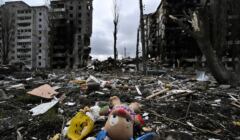 widok ulicy, na ziemi na pierwszym plane leżą dziecięce zabawki, cała ulica zasłana gruzami, stoją 10 piętrowe domy mieszkalne wypalone i częściowo zrujnowane
