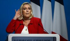 Kobieta, blondynka, w czerwonej marynarce na scenie nadstawia ucha by usłyszeć pytanie z sali. Marine Le Pen