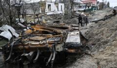 Zniszczony czołg na pierwszym planie, w tle zniszczone domy i dcwoje ludzi idzie resztkami drogi