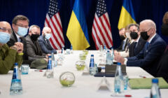 mężczyźni w maseczkach siedzący po obu stronach stołu, w tle flagi amerykańskie i ukraińskie