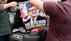 Dwóch mężczyzn wychodzących poza kadr trzyma plakat z Mèlenchonem