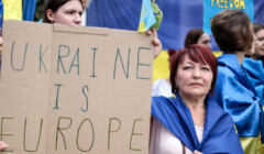 Ludzie trzymają ukrainskie i unijne flagi oraz angielski napis 