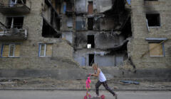 Dziewczynka na hulajnodze przejeżdża obo zbombardowanego domu