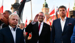 Grzegorz Braun i dwóch innych mężczyzn pod kolumną Zygmunta, w tle flagi