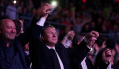 Jacek Kurski ubrany w koszulę i płaszcz, trzyma za ręce inne osoby na widowni podczas koncertu w amfiteatrze