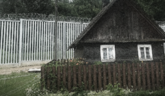 Zdjęcie starej, wiejskiej chaty z ogródkiem, tuż za nią stoi duży metalowy płot z drutem kolczastym