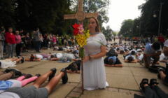 Kobieta w białej sukni klęczy trzymając wielki krzyż, wokół leżą na ziemi pielgrzymi