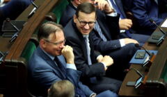 W ławach rządowych w Sejmie siedzi Arkadiusz Mularczyk śmiejący się, obok niego uśmiechnięty Mateusz Morawiecki