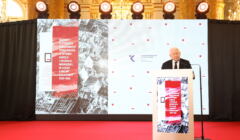 Jarosław Kaczyński na tle ekranu wyświetlający raport o reparacjach wojennych
